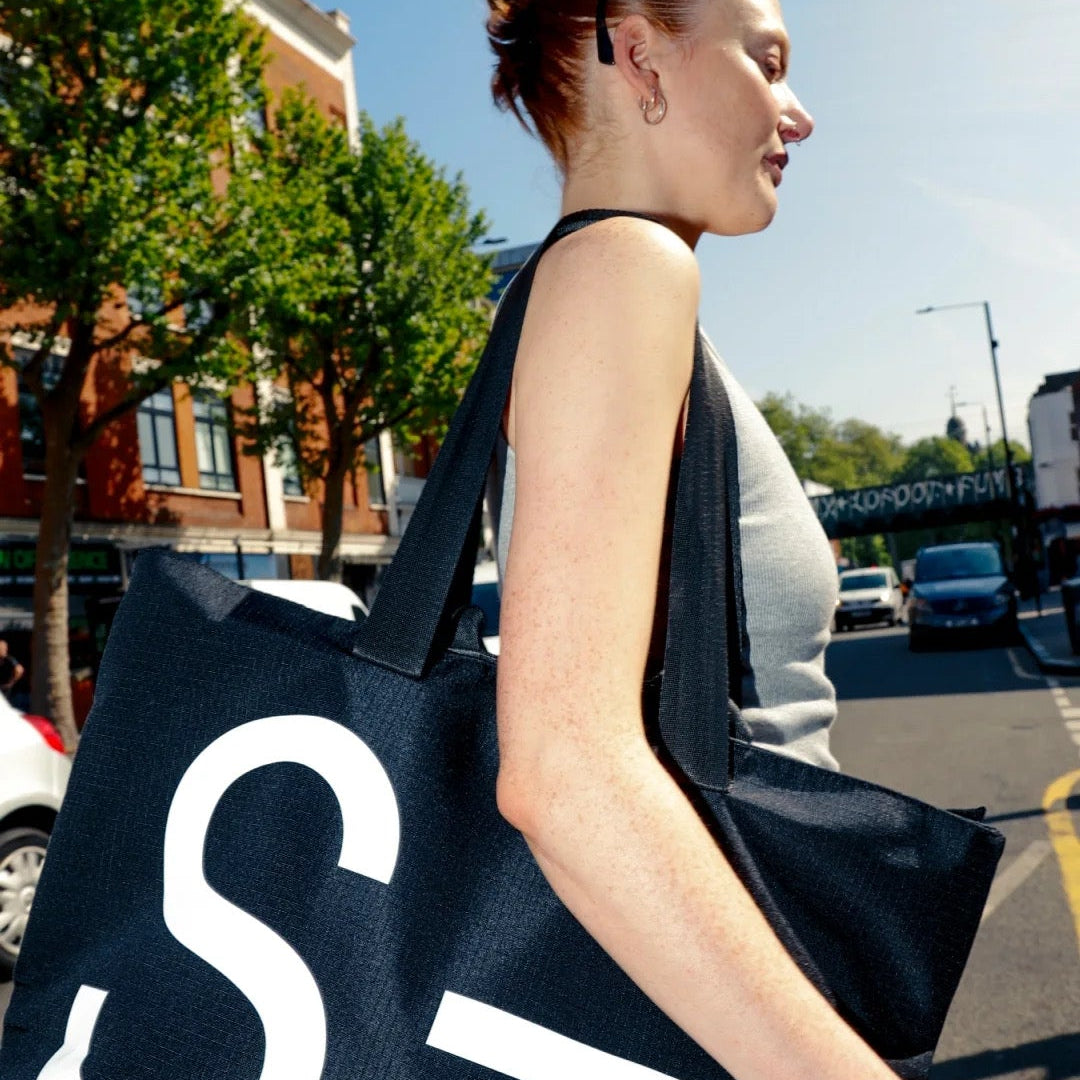 Woman carrying a large black bag with a white 'S' on a city street.