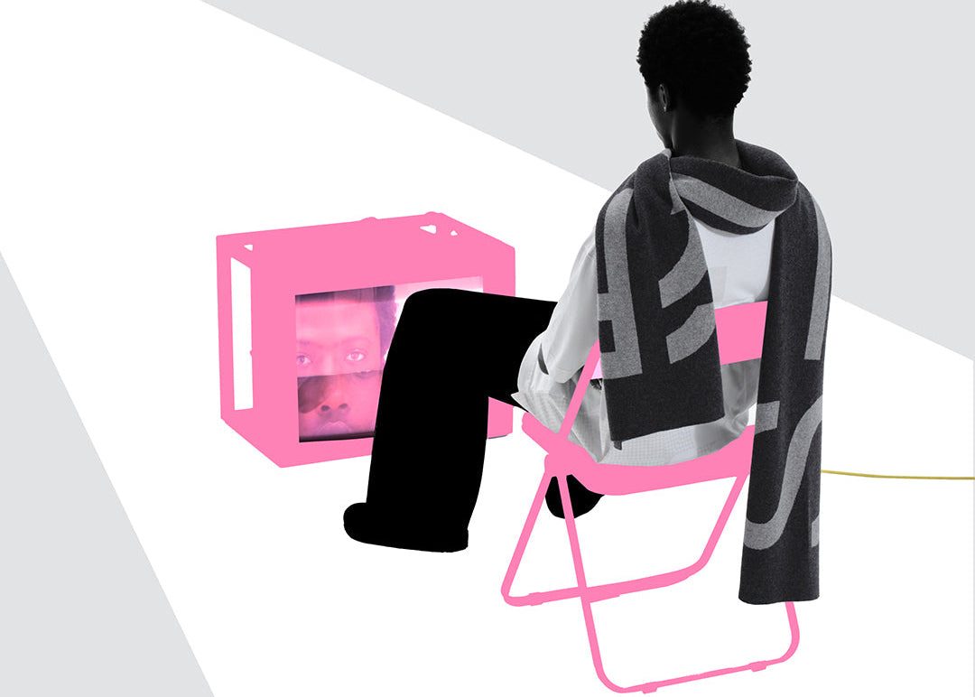 Person sitting on a pink chair with a geometric-patterned blanket, facing a pink computer monitor on a white background