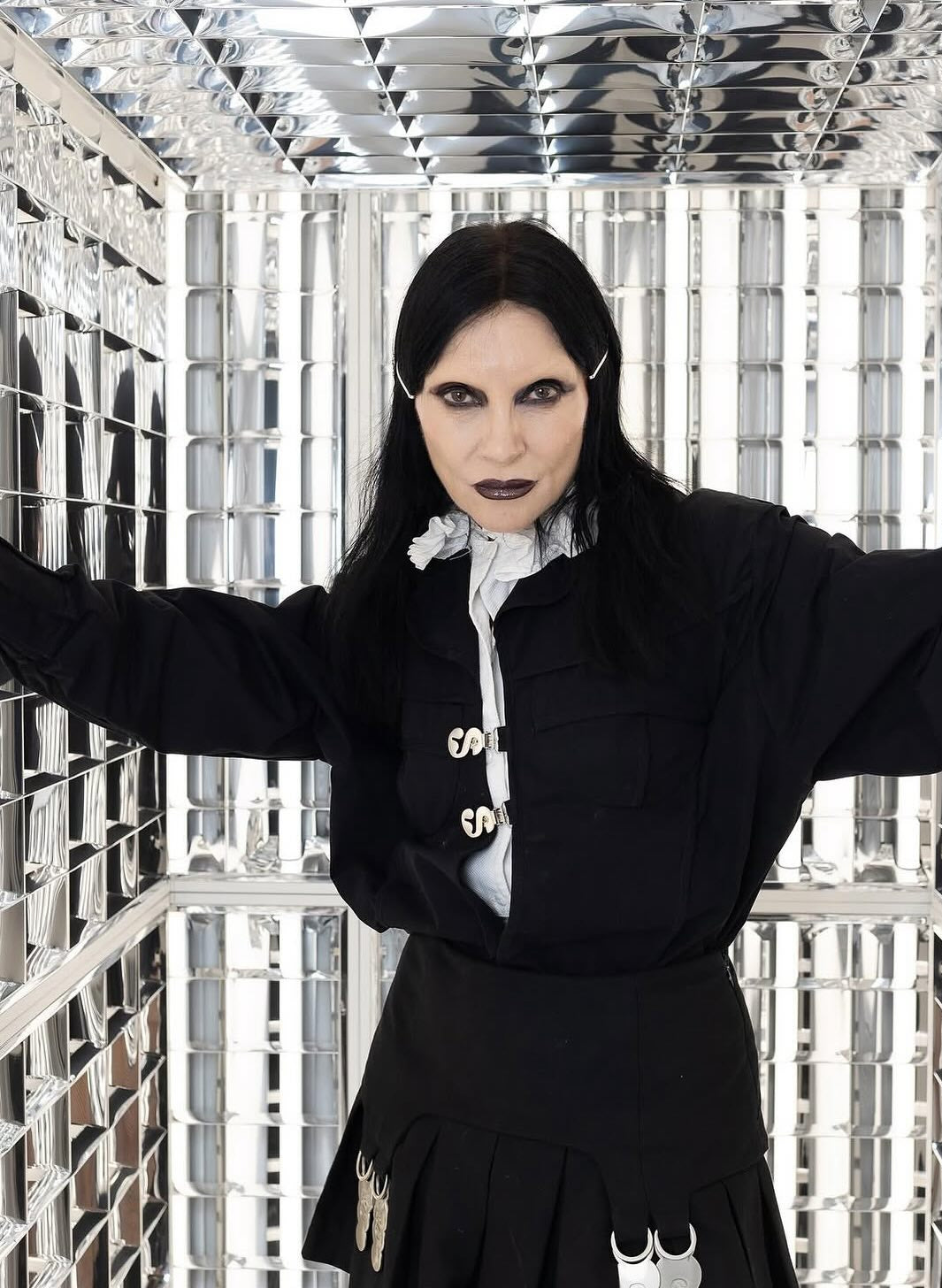 Person in a black outfit with white accents standing in a geometric, reflective room.