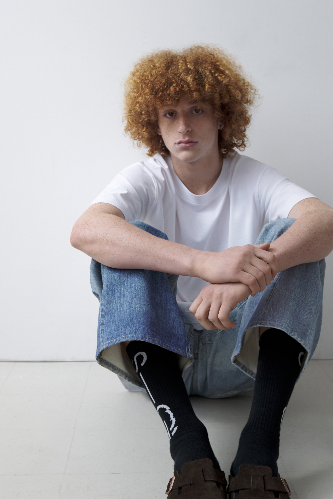 Person with curly hair wearing a white t-shirt, blue jeans, and dark shoes on a white background