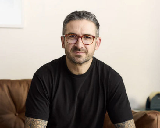 Man with tattoos wearing a black t-shirt and glasses sitting on a brown couch.