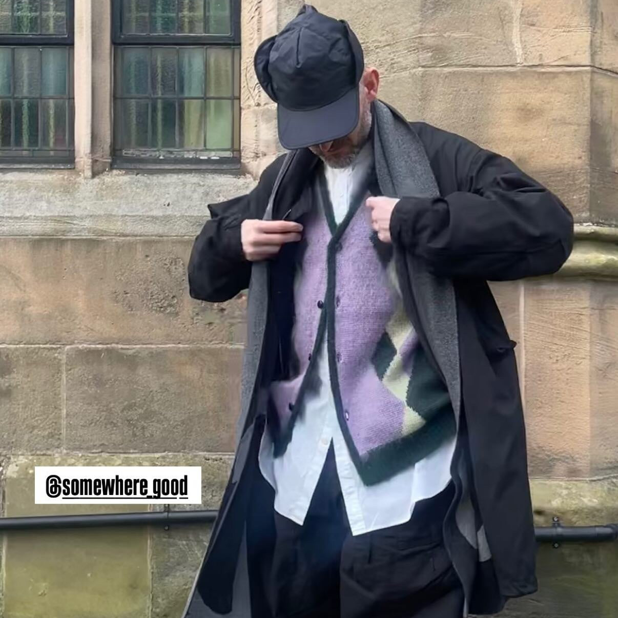 Person wearing a long coat, patterned scarf, and cap in front of a stone building.