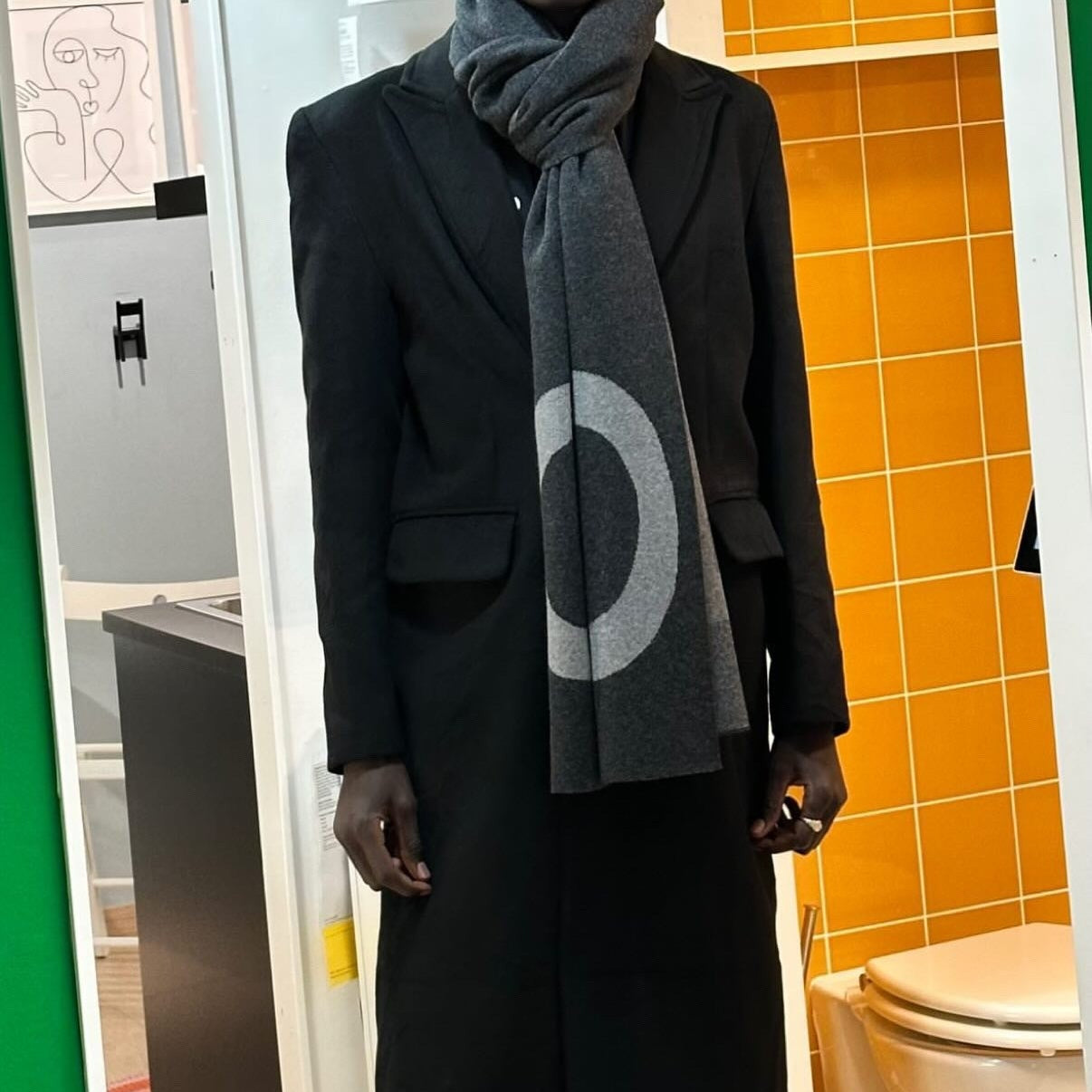Person wearing a long black coat and gray scarf in a bathroom setting