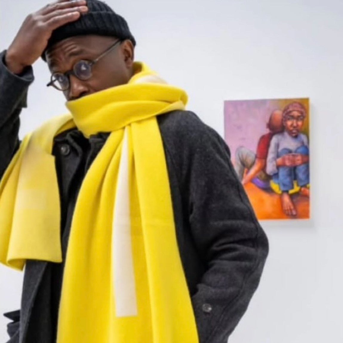 Person wearing a bright yellow scarf with a white logo, standing against a white wall with a colorful painting.